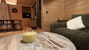 Een gezellige woonkamer met houten wanden, een ronde marmeren salontafel met een brandende kaars, een groene bank met een wit kussen, en een eethoek met een houten tafel en stoelen.