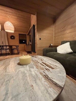 Ein gemütliches Wohnzimmer mit Holzwänden, einem runden Marmortisch mit einer brennenden Kerze, einem grünen Sofa mit einem weißen Zierkissen und einem Essbereich mit Holztisch und Stühlen.