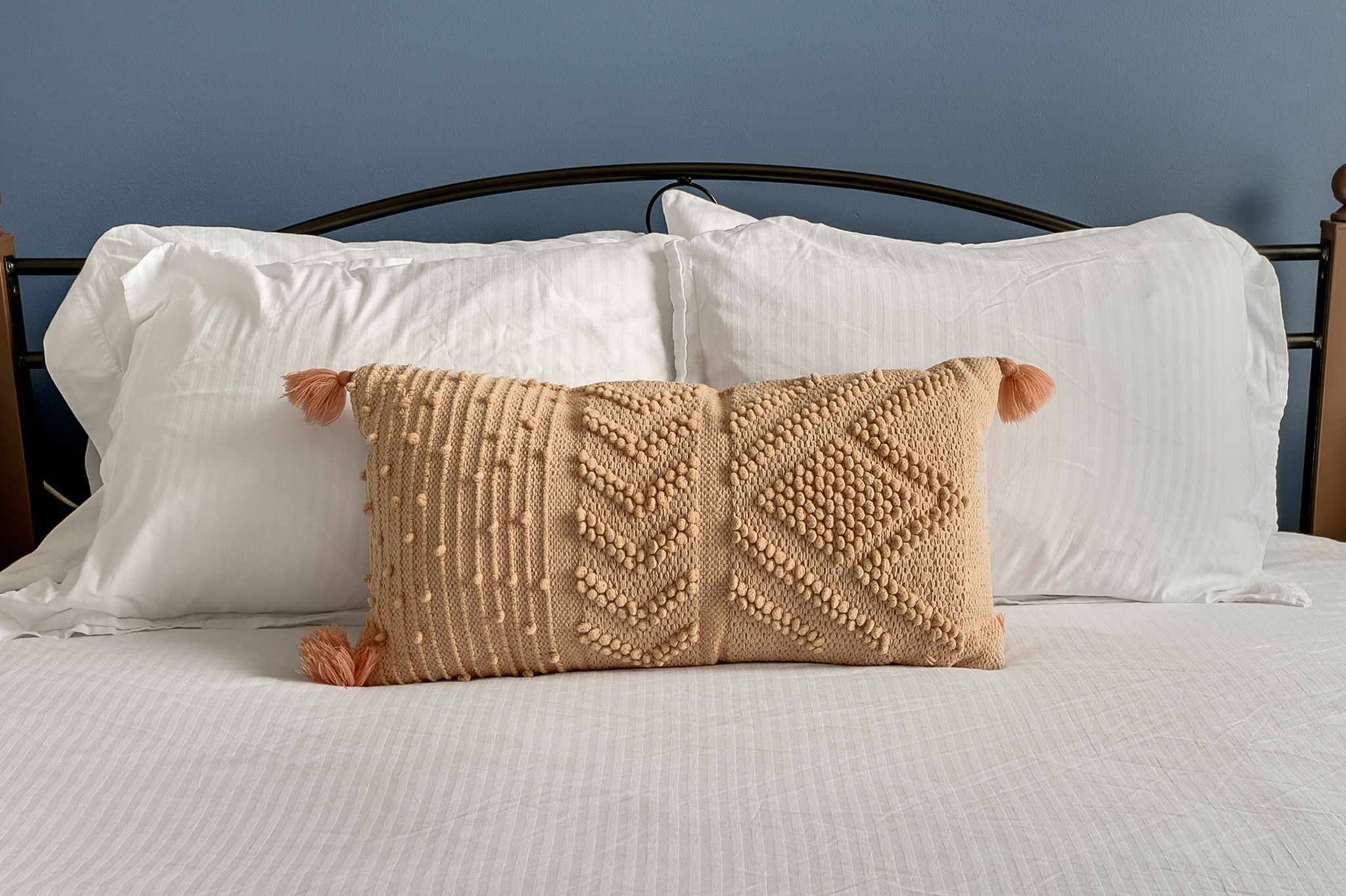 A bed with white linens, a dark metal headboard, and a beige knitted pillow with tassels.