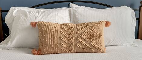 A bed with white linens, a dark metal headboard, and a beige knitted pillow with tassels.