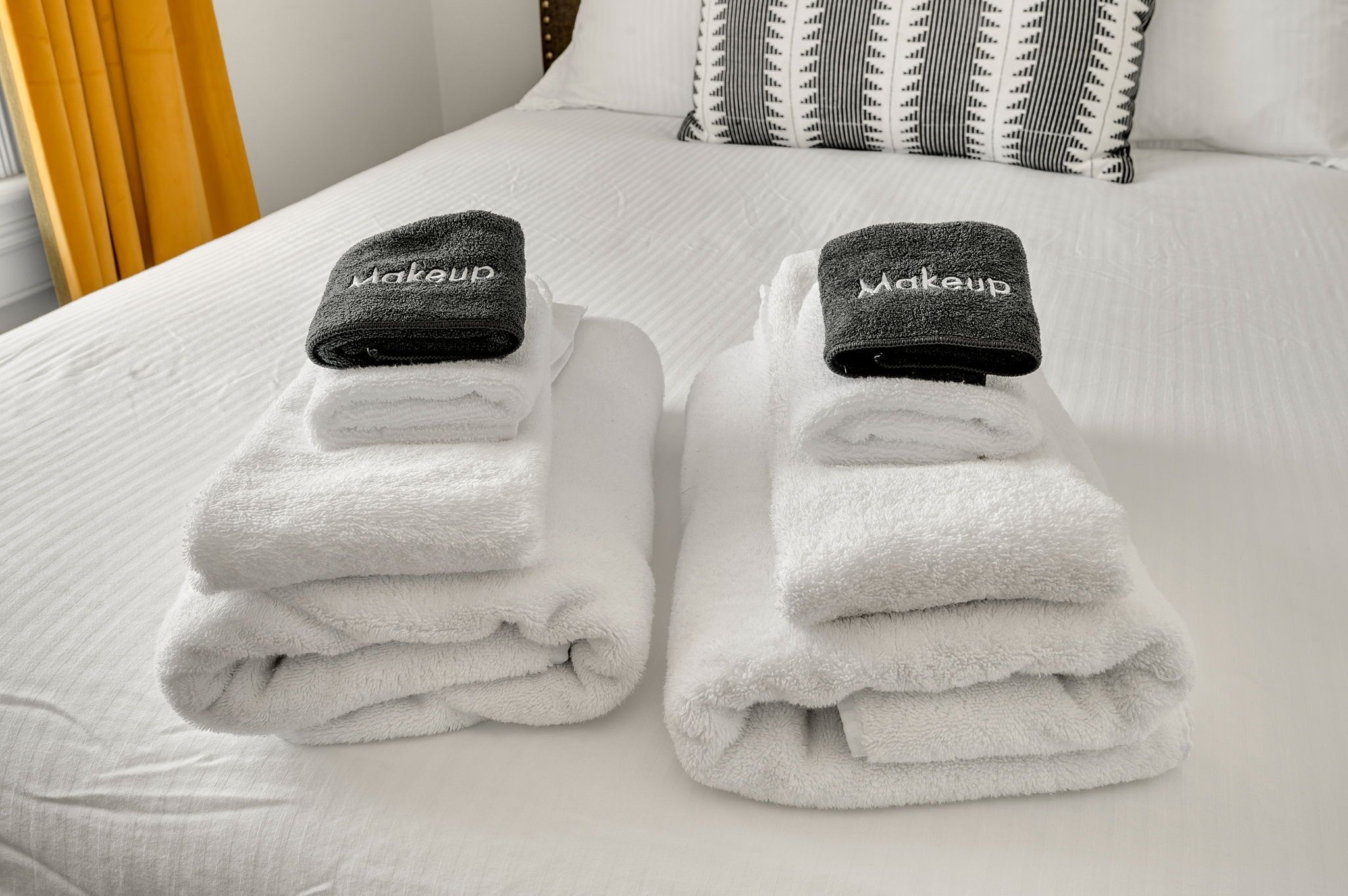 A neatly made bed with white linens and two towels with 'Makeup' branded headbands.