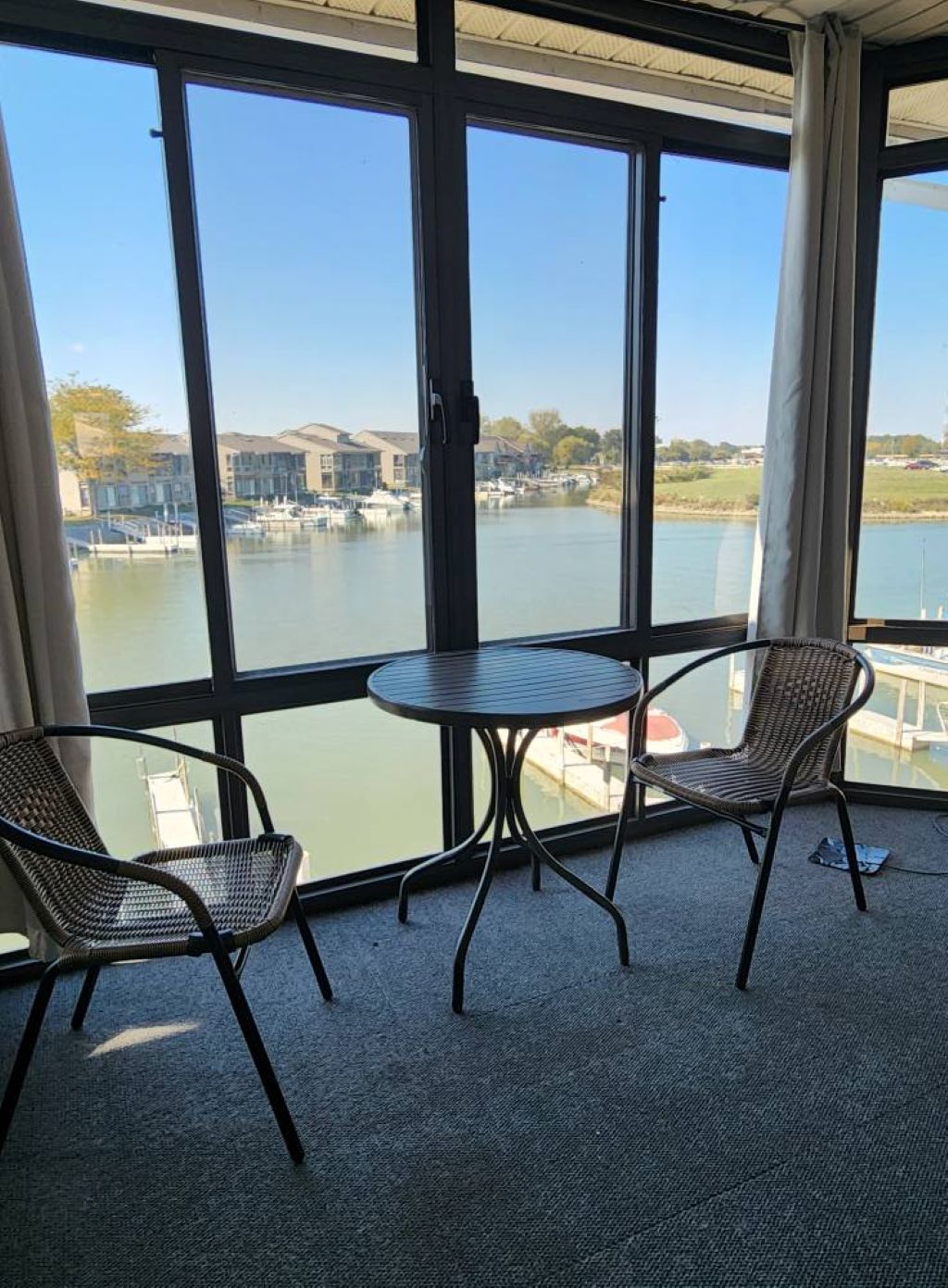 Un balcon avec deux fauteuils en osier et une petite table ronde, donnant sur une marina où l’on peut admirer des bateaux et des bâtiments.