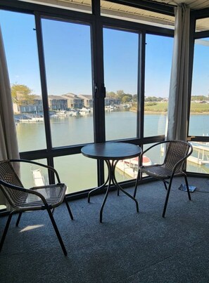 A balcony with two wicker chairs and a small round table, overlooking a marina with boats and buildings.