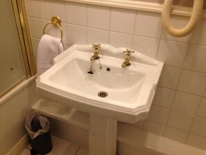 A white pedestal sink with two gold faucets, a towel ring, and a trash bin in a bathroom with white tiled walls. - Ty Newydd Country Hotel (Hirwaun)