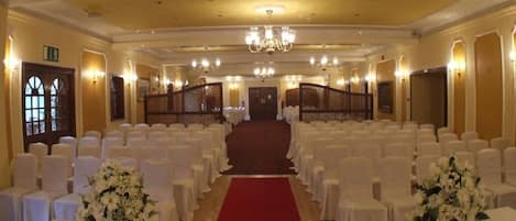 A banquet hall with rows of white chairs, a red carpet aisle, and floral centerpieces.