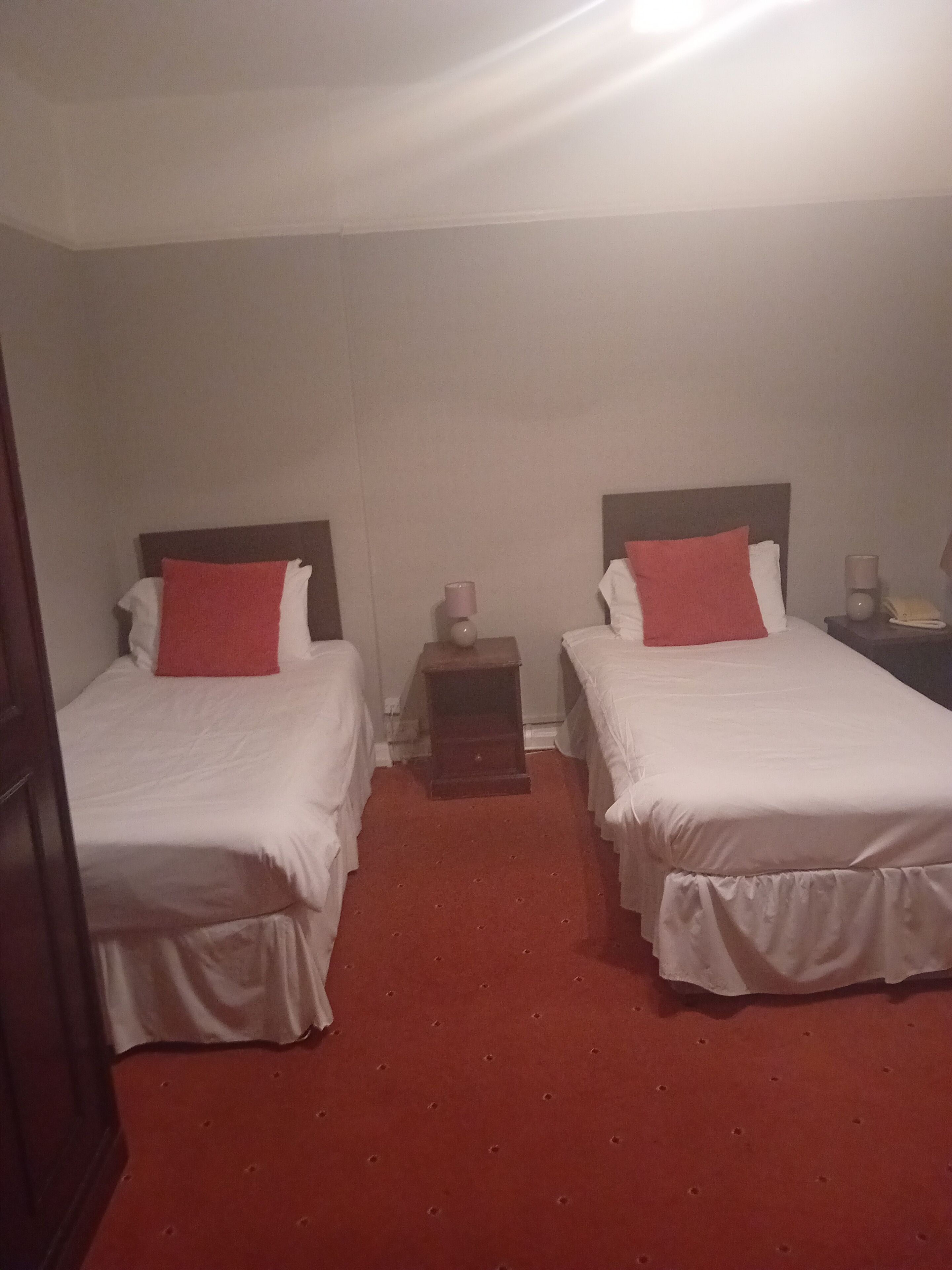 A hotel room with two beds, each with white bedding and red pillows, a small bedside table with a lamp, and a red carpet.