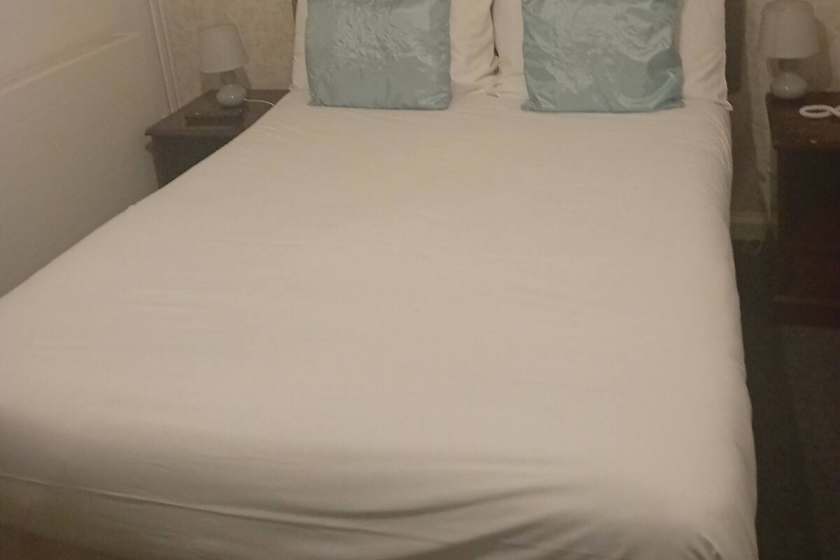 Ett hotellrum med en enkel säng, två kuddar, ett nattduksbord med en lampa och en väggmonterad lampa.