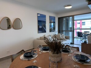 A dining area with a wooden table set for four, a vase with branches, and a view of a building through large windows. - Modern apartment with ocean view and gourmet balcony. (Bertioga)