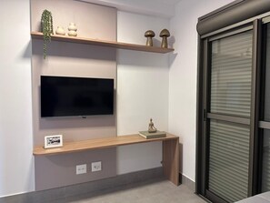 A modern living room with a flat-screen TV mounted on the wall, wooden shelves above, and a small table with decorative items. - Modern apartment with ocean view and gourmet balcony. (Bertioga)