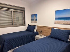 A hotel room with two beds, a window with blinds, and two beach-themed wall art pieces. - Modern apartment with ocean view and gourmet balcony. (Bertioga)
