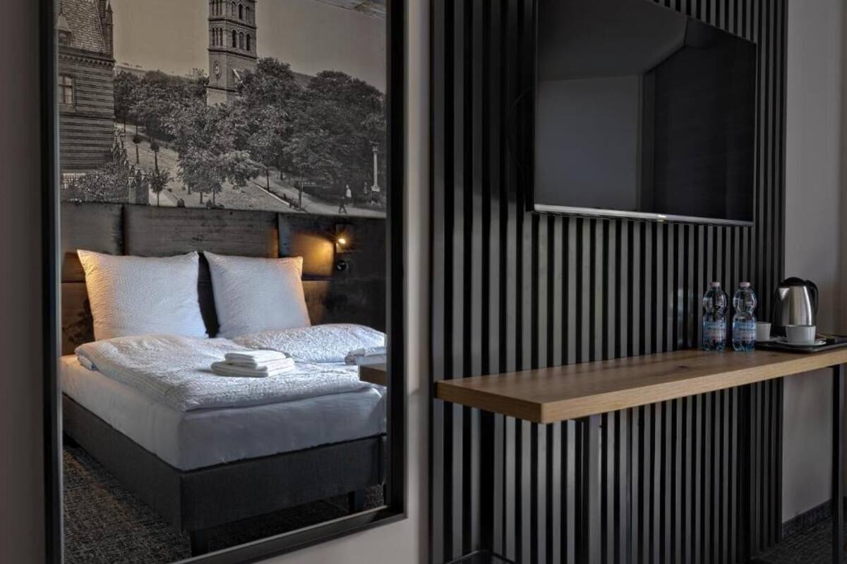A modern hotel room with a large bed, a wooden bedside table, a flat-screen TV, and a framed cityscape artwork on the wall.