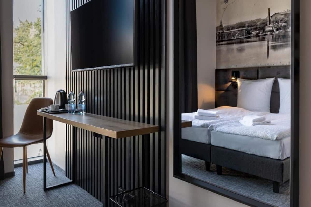 A modern hotel room with a bed, a desk with a chair, a TV, and a window with a view of a cityscape.