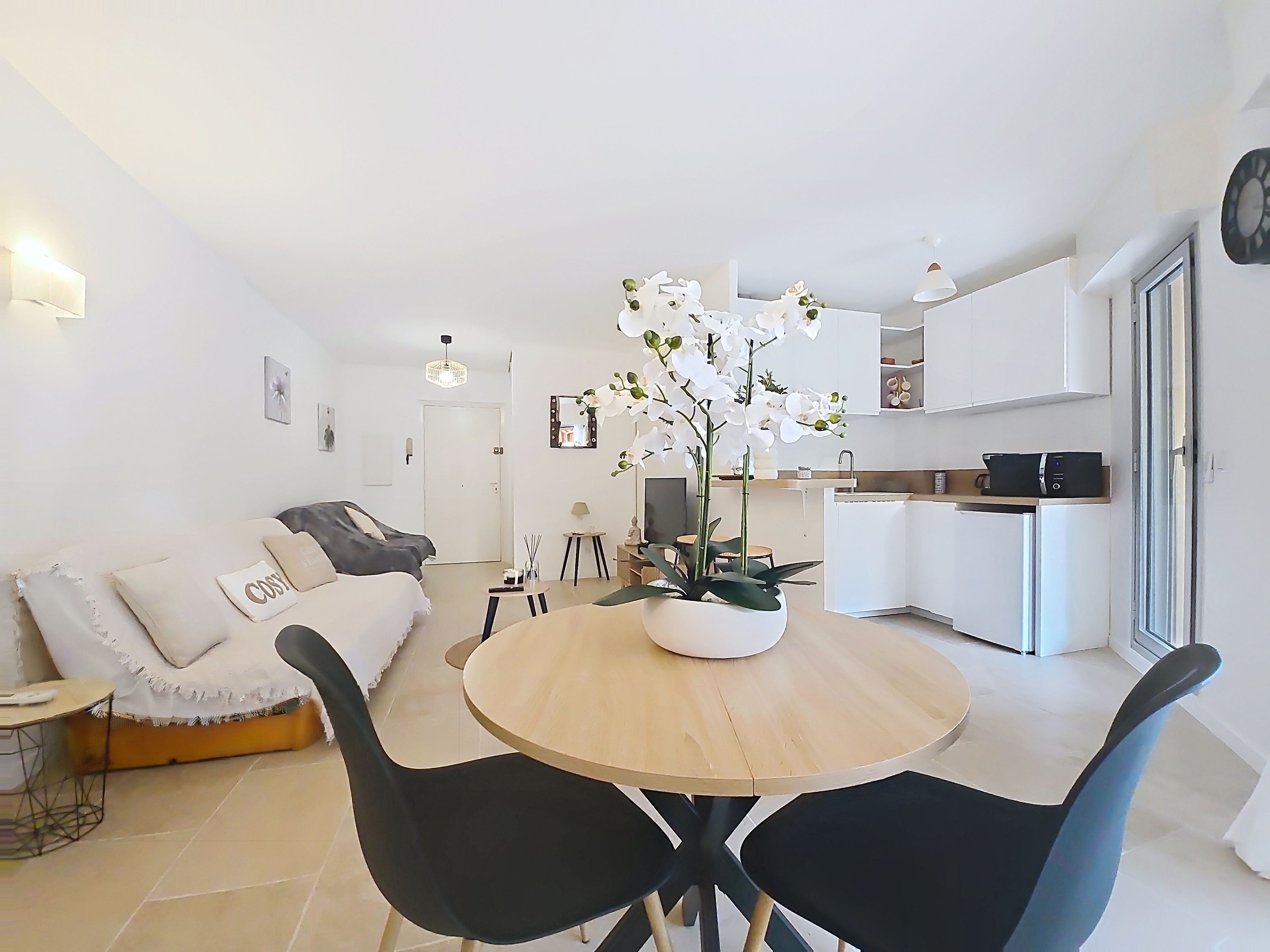 A modern living room with a sofa, a round table, and chairs. There is a kitchen area with cabinets and a sink.