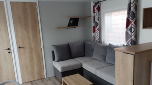 Ett modernt vardagsrum med en grå hörnsoffa, ett soffbord i trä, en liten hylla och ett fönster med mönstrade gardiner.