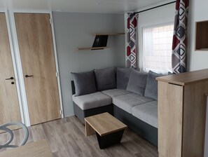 Un salon moderne avec un canapé d’angle gris, une table basse en bois, une petite étagère et une fenêtre agrémentée de rideaux à motifs.
