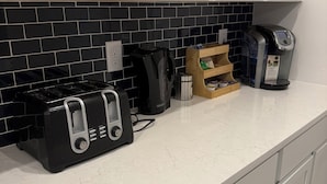 Ett kök med vita skåp, en svart kakelbeklädd vägg bakom arbetsbänken, en kaffebryggare, en brödrost och en vattenkokare.