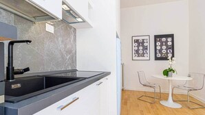 Een moderne keuken met een zwart aanrecht en witte kasten, een eethoek met een witte tafel en transparante stoelen, en wanddecoratie met ingelijste foto's.