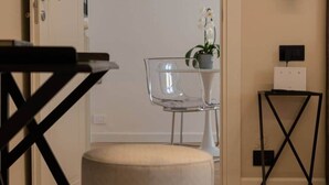 Een minimalistisch ingerichte kamer met een transparante glazen stoel, een klein rond voetenbankje, een zwarte metalen tafel en een orchidee in een pot op een plank.