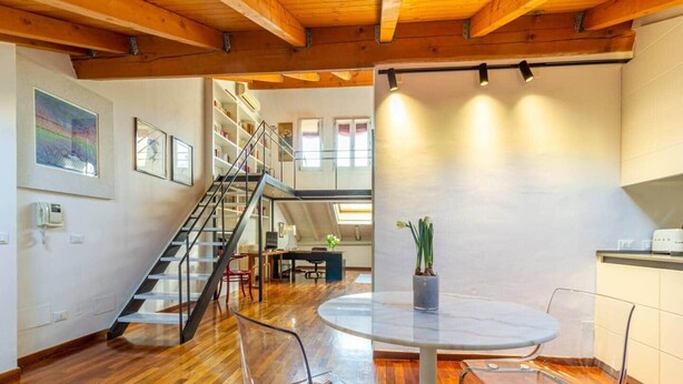 A spacious room with a wooden floor, a round table with chairs, a staircase, and a kitchen area. - Casa Pegaso in Canton de Fiori (Bologna)