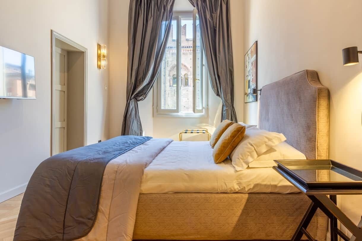 Une chambre d’hôtel avec un lit, une table de chevet, une fenêtre avec des rideaux, un téléviseur fixé au mur et un tableau encadré.