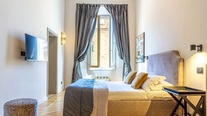 Een slaapkamer met een bed, een televisie, een raam met gordijnen, een lamp en een klein tafeltje.