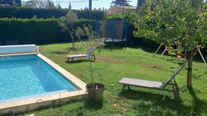 Piscina retangular com borda azulejada, rodeada por grama e um viveiro. Há duas espreguiçadeiras e uma pequena mesa perto da piscina.