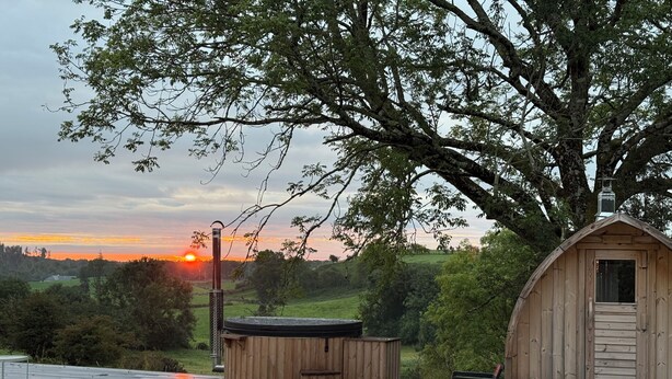Eine Holzhütte mit Schornstein, einem Whirlpool und einem Stuhl auf einer gefliesten Terrasse mit Blick auf einen Sonnenuntergang.