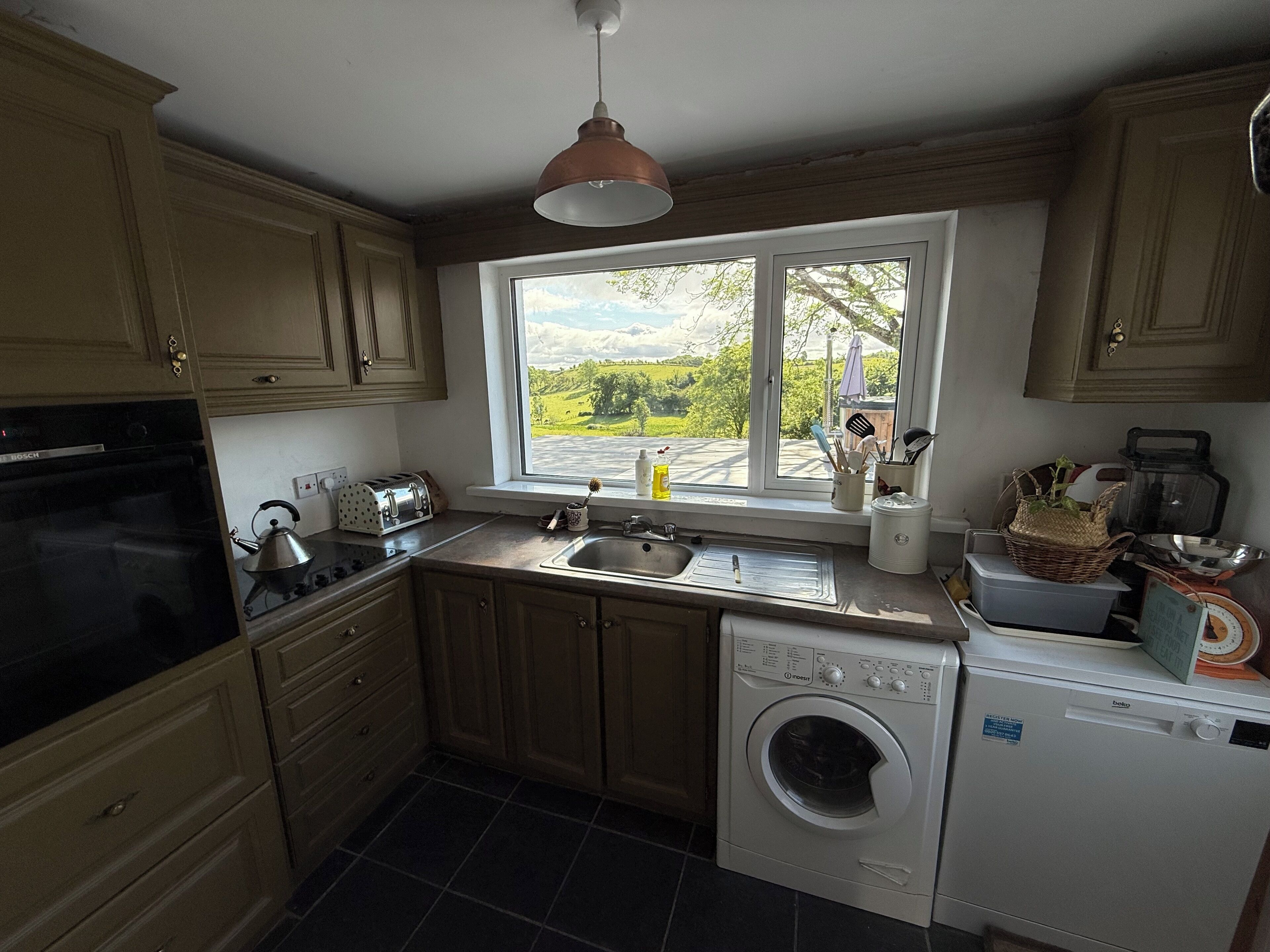 Une cuisine avec des meubles en bois, un lave-linge Bosch, un lave-vaisselle Bosch, une fenêtre donnant sur un espace verdoyant et un luminaire suspendu.
