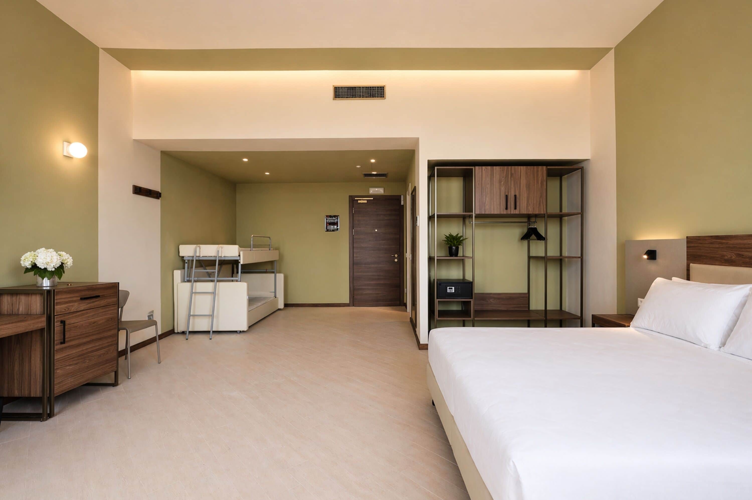 A hotel room with a bed, desk, chair, wardrobe, and a small kitchenette area.