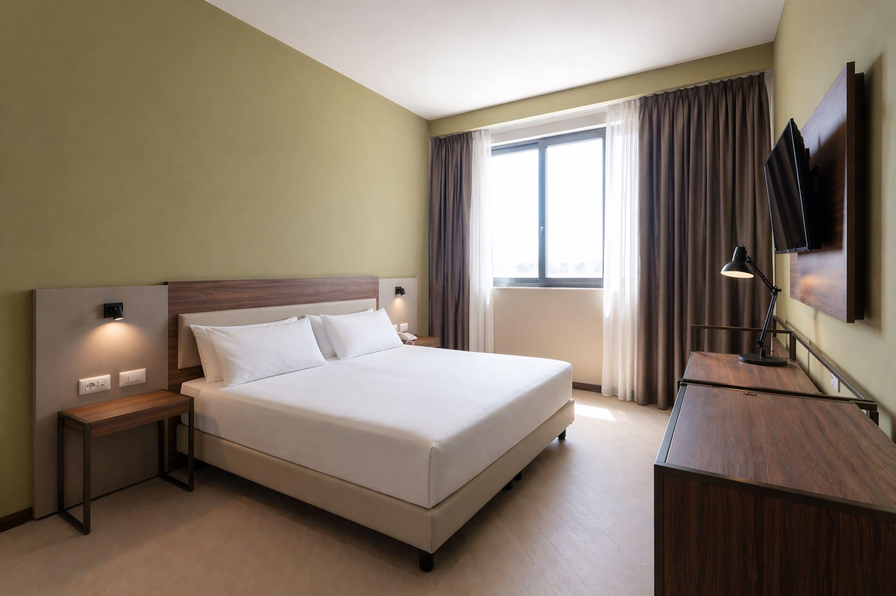A hotel room with a bed, bedside table, lamp, TV, and a large window with curtains.