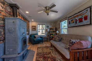 A cozy living room with a stone fireplace, a ceiling fan, a sofa with patterned cushions, a bookshelf, and a large window.