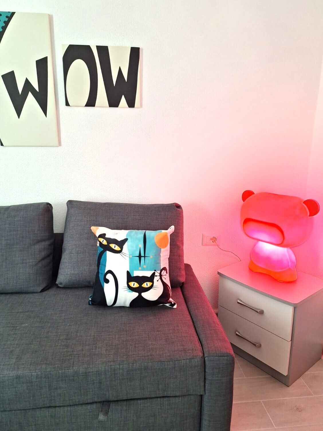 Una sala moderna con un sofá gris, un cojín decorativo, una mesita de noche con cajón y una pared con un letrero que dice 'WOW'.