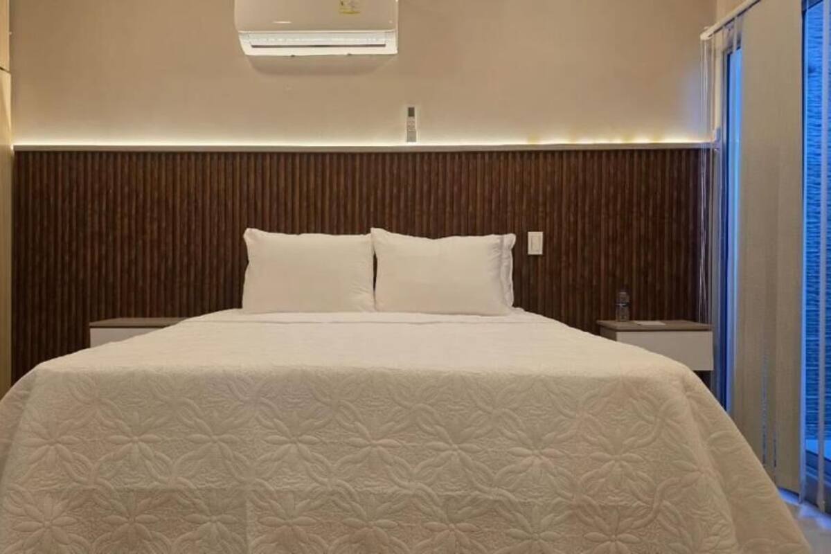 Een slaapkamer met een groot bed, een houten hoofdbord, nachtkastjes en een raam met jaloezieën.
