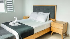 A neatly made bed with a grey headboard, white bedding, and a grey bench at the foot of the bed. There is a wooden bedside table with drawers and a small nightstand with a drawer. - Lyra Vacation Rentals (Willemstad)