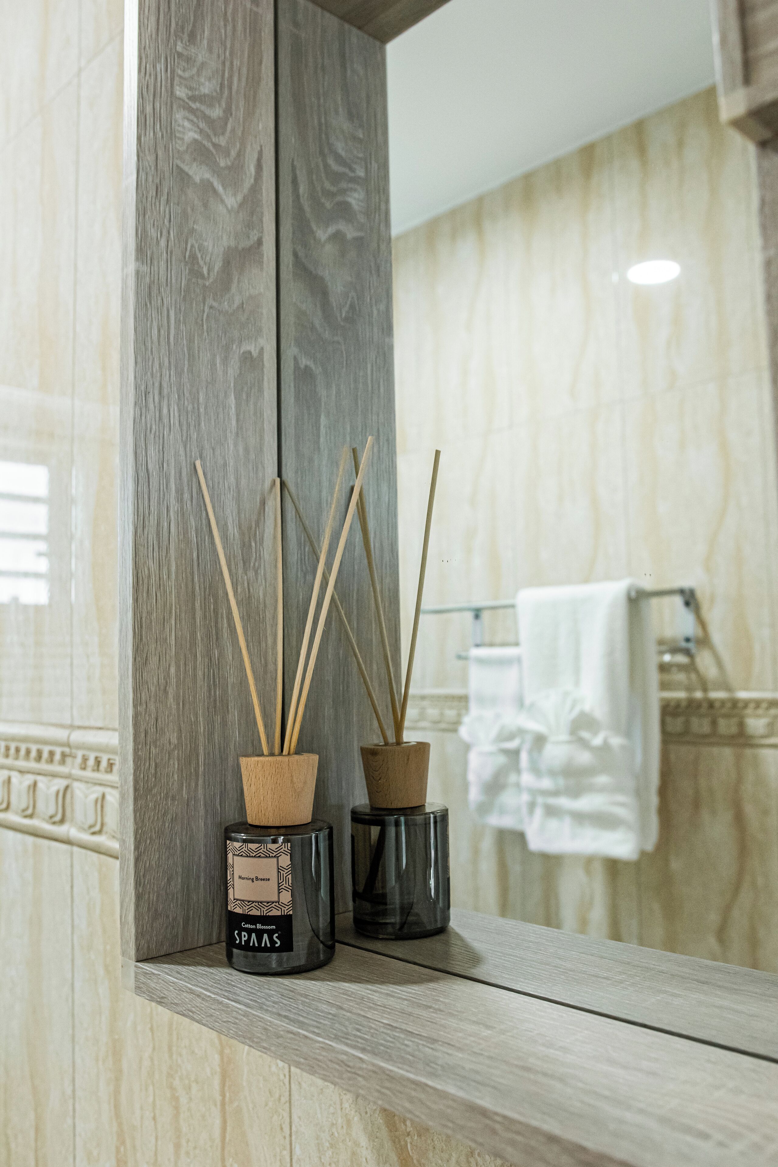A bathroom with a wooden shelf holding two reed diffusers and a towel rack with a white towel.