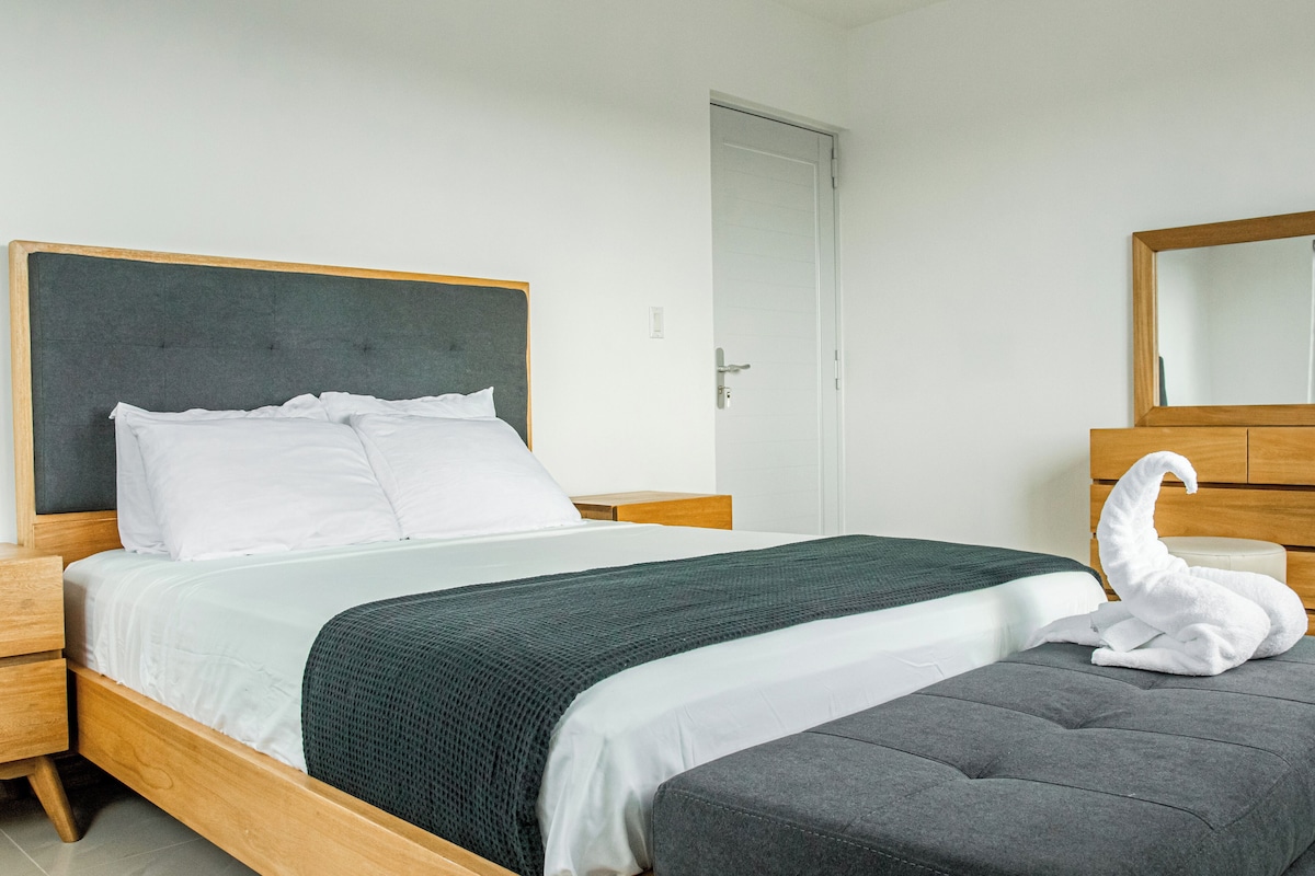 A neatly made bed with white linens and a dark green blanket, a wooden headboard, a grey upholstered bench, and a white swan decoration.