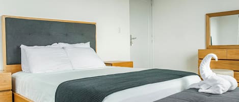 Una cama bien tendida con sábanas blancas y una manta verde oscuro, un cabecero de madera, un banco tapizado de color gris y una decoración con forma de cisne blanco.