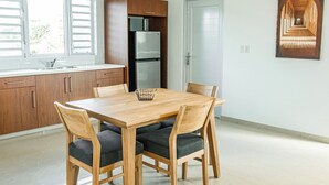A modern kitchen with wooden cabinets, a stainless steel refrigerator, a dining table with chairs, and a window with blinds. - Lyra Vacation Rentals (Willemstad)