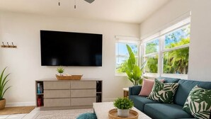 Ein Wohnzimmer mit Flachbildfernseher, Deckenventilator, Sofa, Couchtisch und einem großen Fenster mit Blick auf Grünflächen.