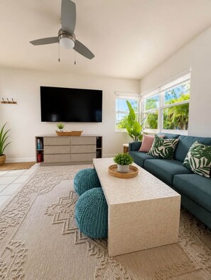 A living room with a flat-screen TV, a ceiling fan, a sofa, a coffee table, and a large window with a view of greenery. - The Emerald Palms Tropical Beach Villa (Hollywood)