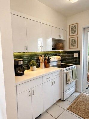 A modern kitchen with white cabinets, a green tiled backsplash, a coffee maker, a stove, and a small plant. - The Emerald Palms Tropical Beach Villa (Hollywood)
