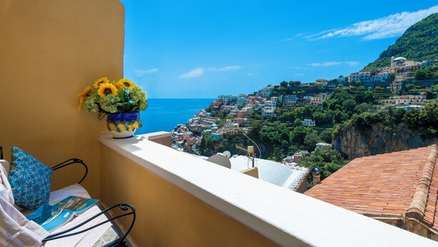 A balcony with a view of a coastal town, a vase of sunflowers, and a patterned cushion. - Barbera House (Positano)