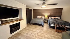 A modern bedroom with a flat-screen TV mounted on the wall, a ceiling fan, two beds with headboards, a wooden floor, and a small seating area.