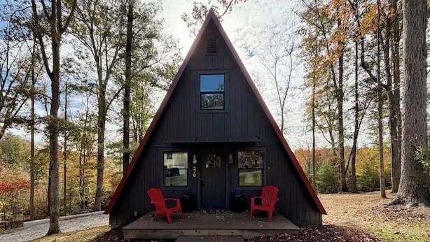 A black triangular cabin with two red Adirondack chairs on a wooden deck, surrounded by trees.