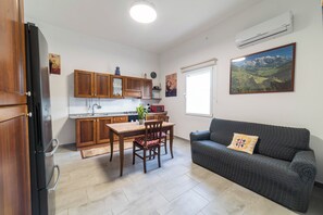 Eine kleine Küche mit Holzschränken, einem Esstisch mit Stühlen, einem grauen Sofa mit Kissen und einem großen Fenster.