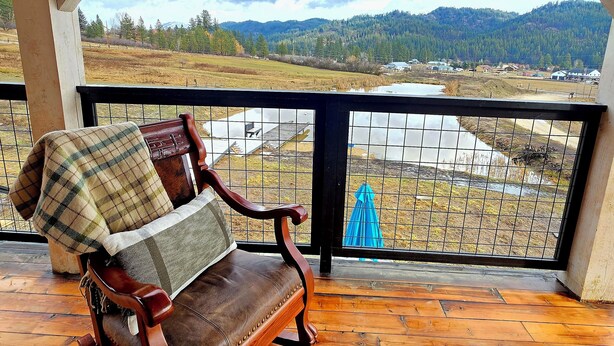 Un banco de madera con una manta escocesa y un cojín, con vistas a un paisaje pintoresco que incluye montañas y un toldo azul en la barandilla.