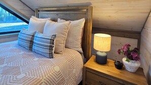 Un dormitorio con techo de madera, una cama con ropa de cama estampada, una mesita de noche con lámpara y un florero con flores.