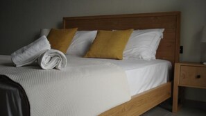 Una cama bien tendida con almohadas blancas y amarillas, una toalla blanca doblada y un cabecero de madera.