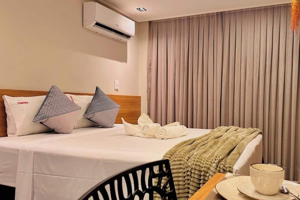 Una habitación de hotel con cama, almohadas, una manta, televisión y ventana con cortinas.
