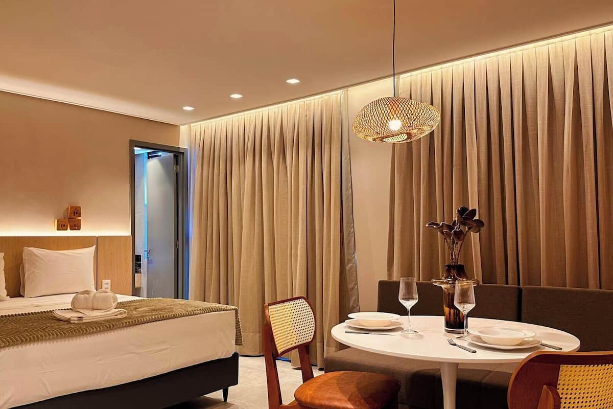 Habitación de hotel moderna con una cama bien hecha, una mesa de comedor para dos personas y un ventanal con cortinas.